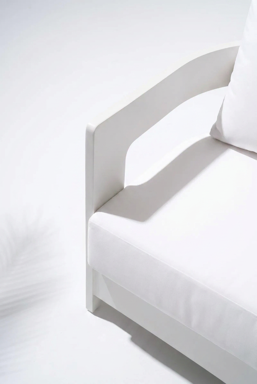A close-up of a modern white upholstered armchair with a curved backrest and a white pillow, photographed in bright studio lighting against a white background.