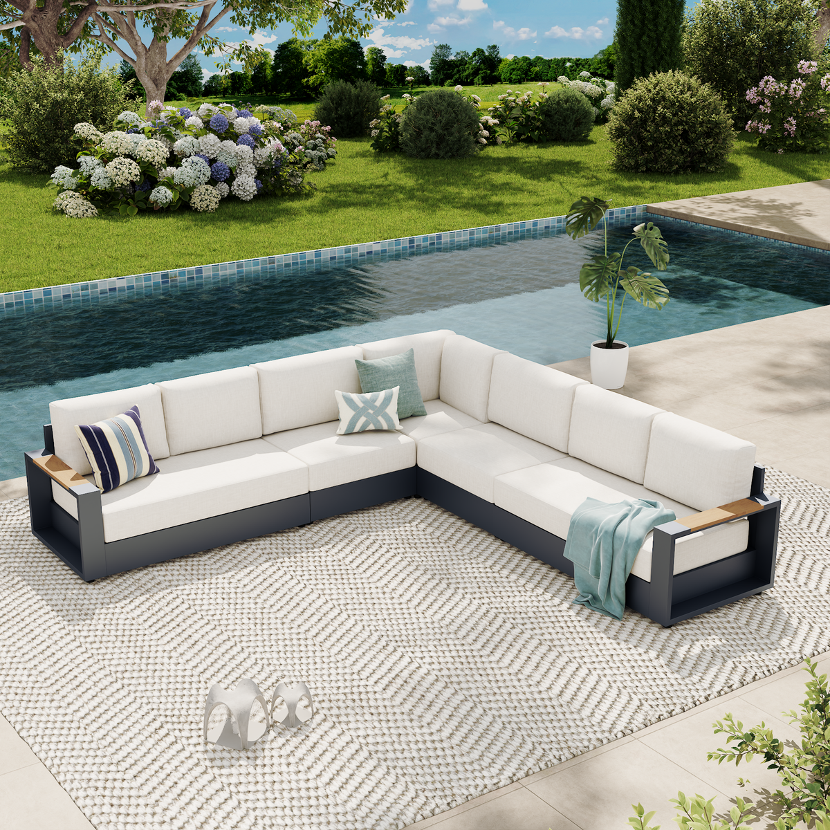 An outdoor sectional sofa set with white cushions and a dark gray frame, arranged on a patterned rug beside a swimming pool. The set includes accent pillows and a light blue throw blanket, with a green lawn, trees, and shrubs in the background. #config_7-Seat L‑Corner