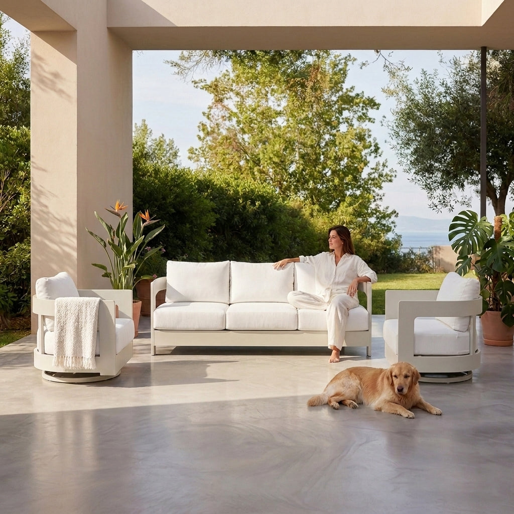 A woman in a white outfit sits on a white outdoor sofa on a covered patio, with two matching white armchairs and a golden retriever lying on the concrete floor. Lush green trees and plants are visible in the background. #color_white