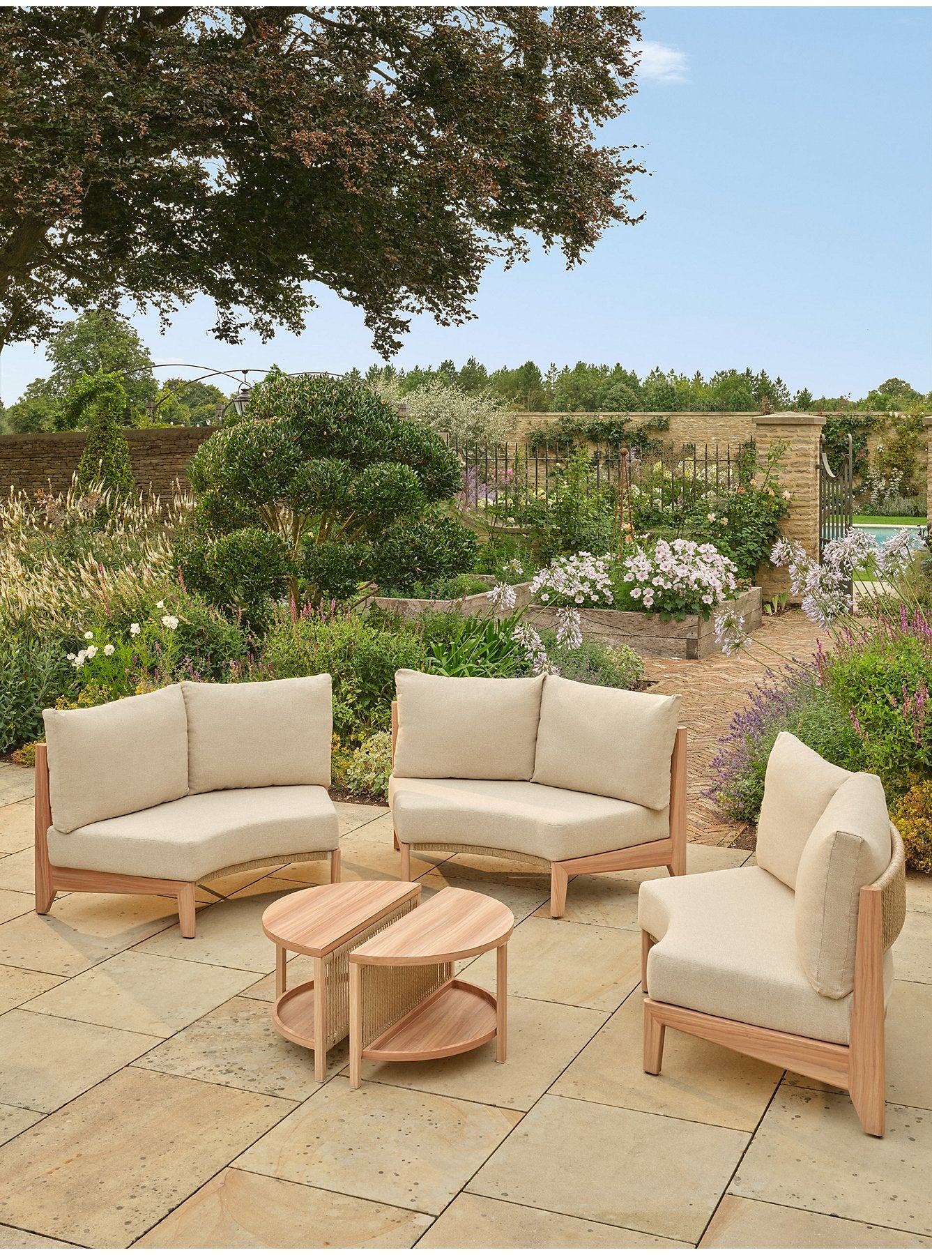 Outdoor patio conversation set with three light beige upholstered armless sofas and two round coffee tables on a stone patio, surrounded by a lush garden- Jardina outdoor modern premium patio furniture