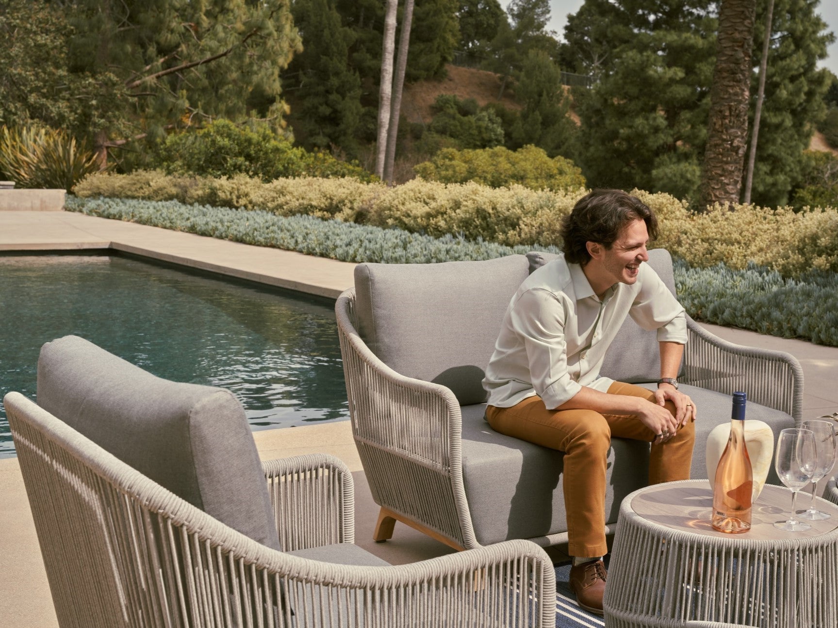 Man sitting on a patio chair by a pool with outdoor furniture and plants in the background