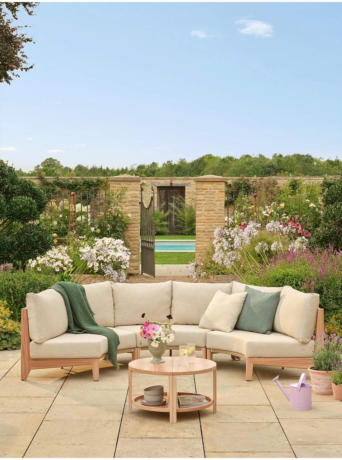 Outdoor sectional patio furniture set with light beige cushions on a stone patio in a garden setting with a round wooden coffee table and decorative pillows- Jardina outdoor modern premium patio furniture